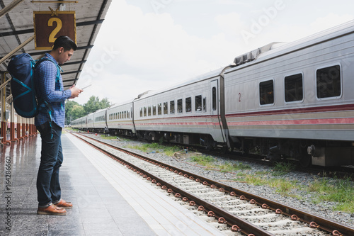 asian man with backpack standing on platform at train station. backpacker or traveler look at digital tablet while waiting for train. journey, trip, travel concept © 88studio