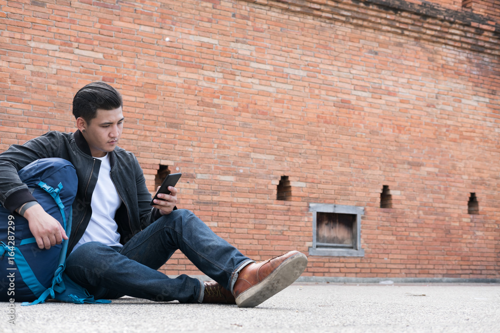 young traveler, asian man wearing black jacket and blue jeans sitting near old orange brick wall with mobile smart phone and backpack young traveler, asian man wearing black jacket and blue jeans sitting near old orange brick wall with mobile smart phone and backpack