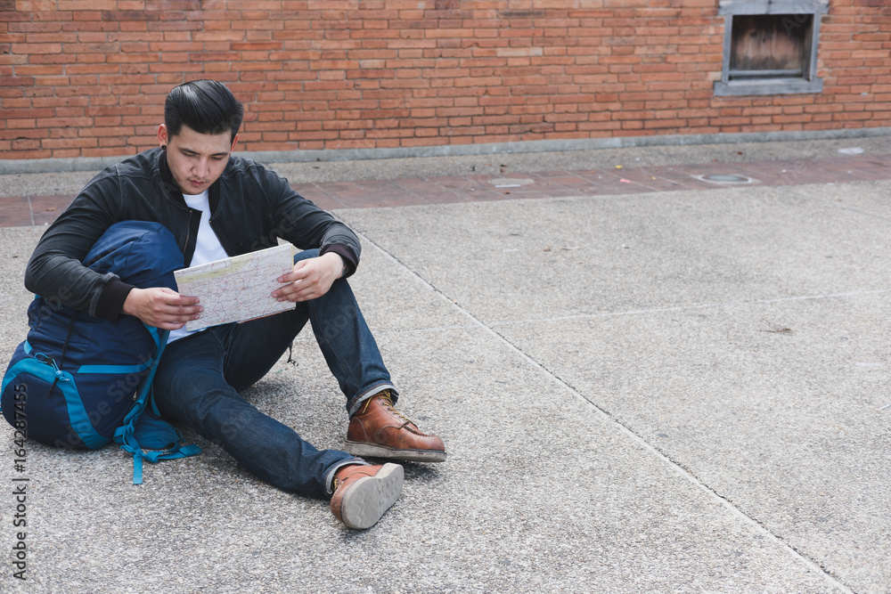 young traveler reading map. asian man wearing black jacket and blue jeans sitting near old orange brick wall with backpack