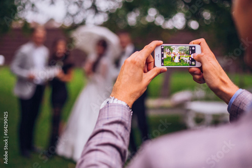 Obraz na płótnie Man taking photo on phone of stylish wedding bride and groom posing. photo booth. wedding couple making photos with friends on phone camera. hand holding smartphone w salonie