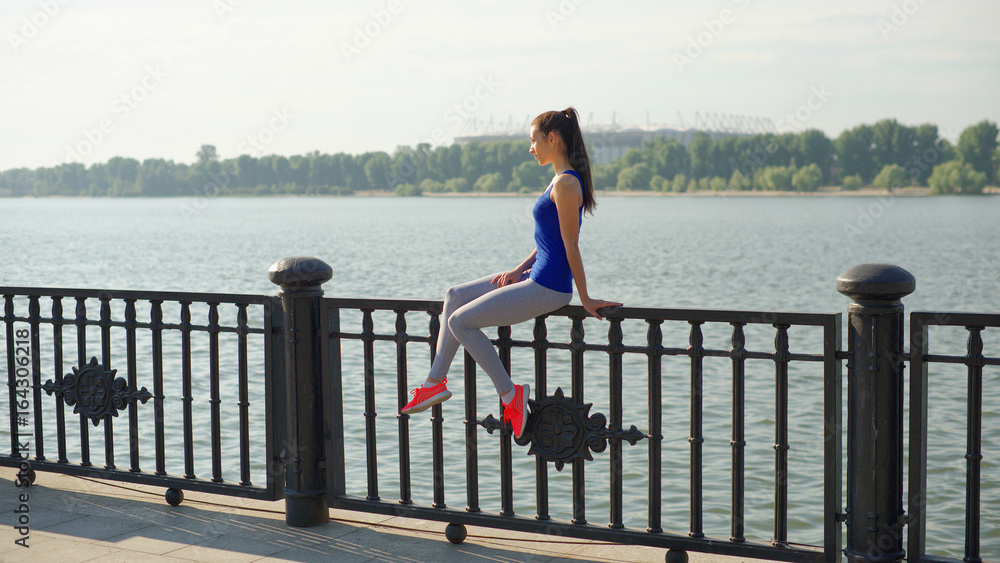Young sporty woman resting while sitting on the railing on the waterfront
