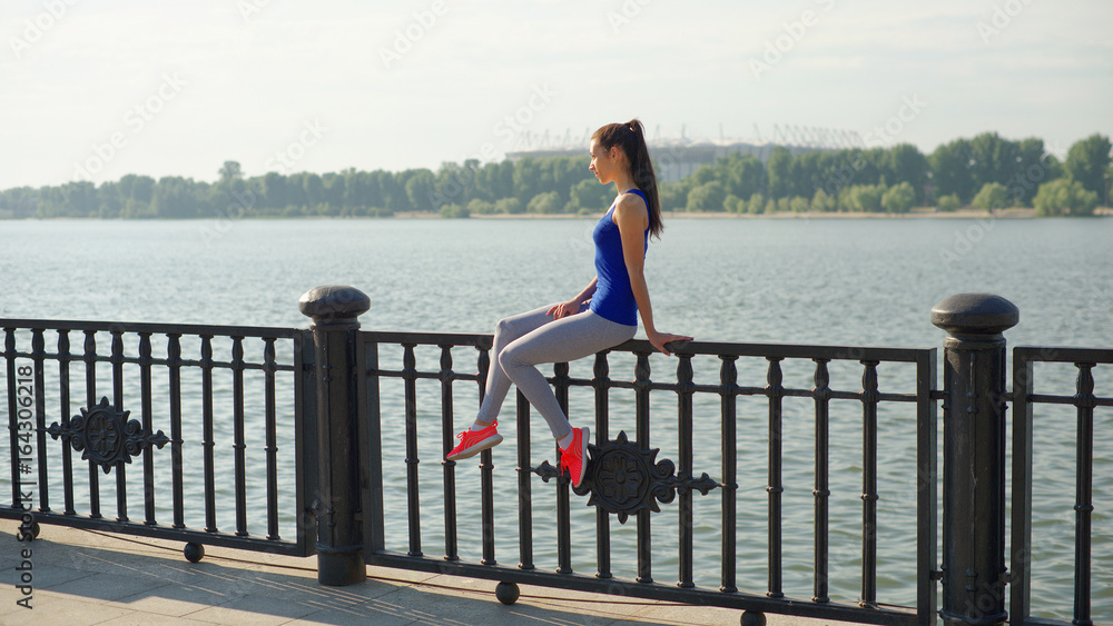 Young sporty woman resting while sitting on the railing on the waterfront Young sporty woman resting while sitting on the railing on the waterfront