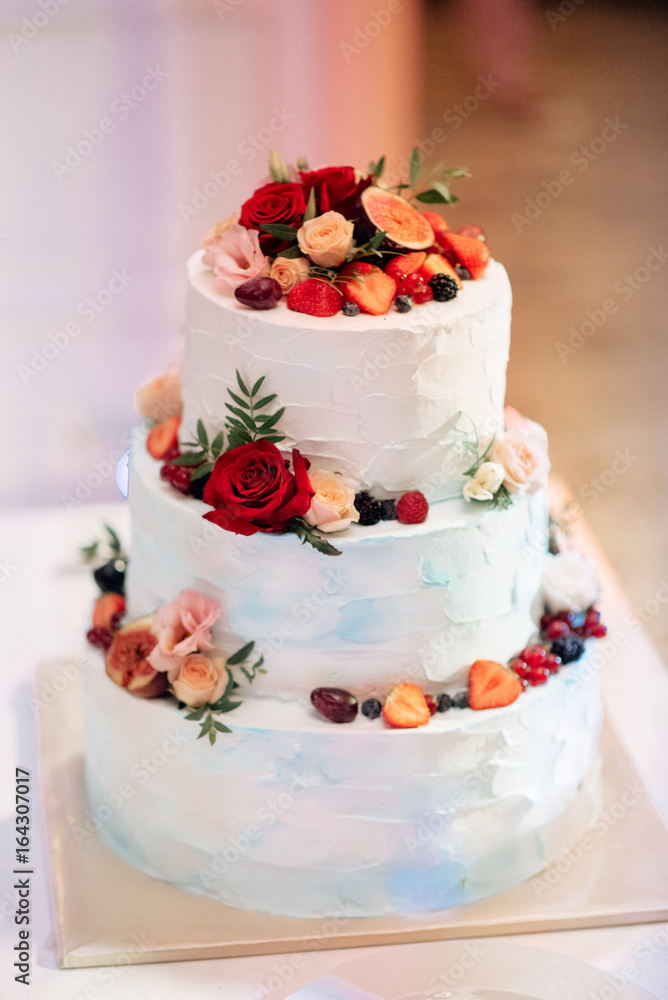 White wedding cake decorated with red, pink roses and fruits, standing on golden plate White wedding cake decorated with red, pink roses and fruits, standing on golden plate