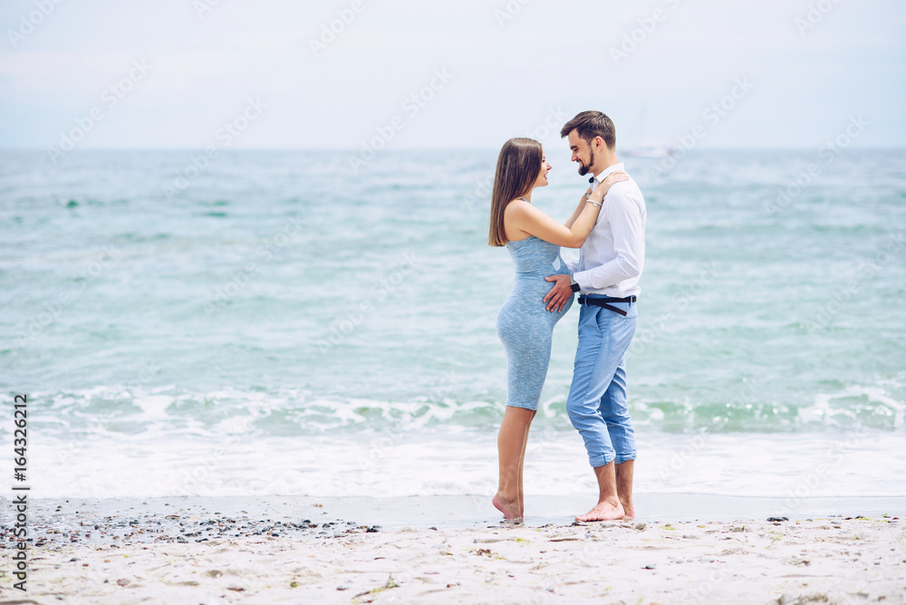 A beautiful smiling pregnant woman in a blue dress and her courageous and handsome husband in a shirt and blue trousers leaned to each other against the background of the sea.