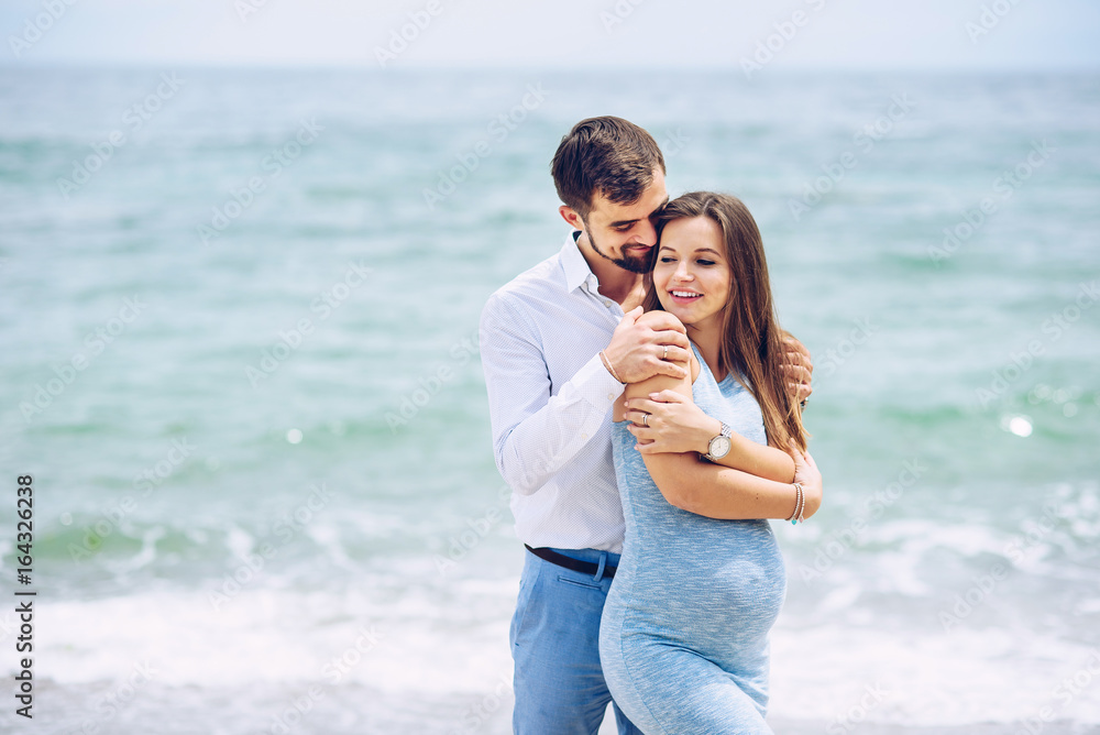 Courageous and handsome man in a white shirt and blue trousers hugs her beautiful pregnant wife in a blue dress against the background of the sea. Courageous and handsome man in a white shirt and blue trousers hugs her beautiful pregnant wife in a blue dress against the background of the sea.