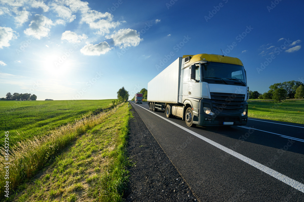 Trucks driving on asphalt road along the green fields in rural landscape at sunset