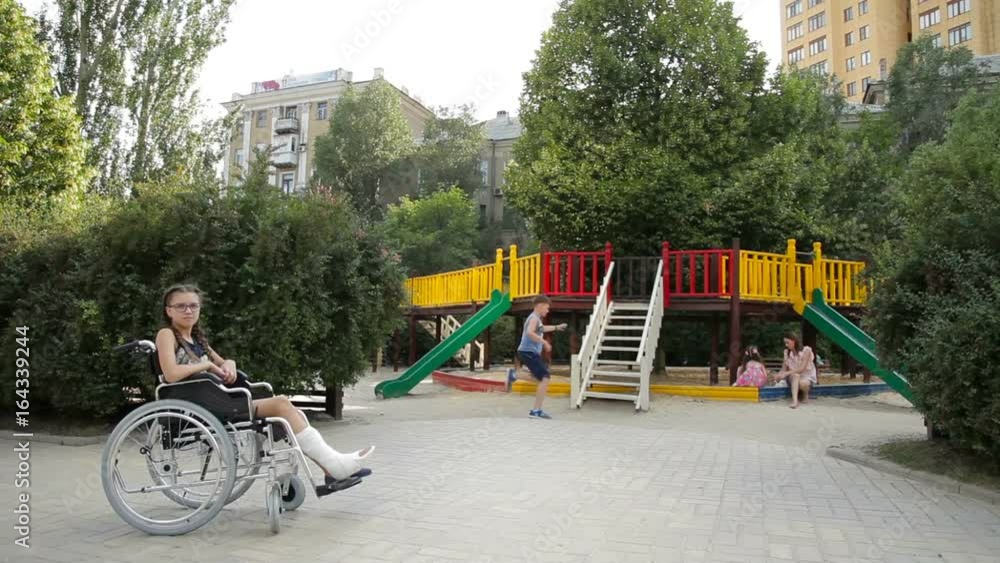 A girl with a broken leg sits in a wheelchair in front of the playground