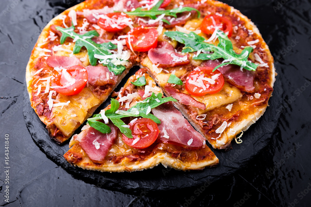 Homemade pizza with prosciutto, tomato, arugula on black slate board. Close up. Homemade pizza with prosciutto, tomato, arugula on black slate board. Close up.