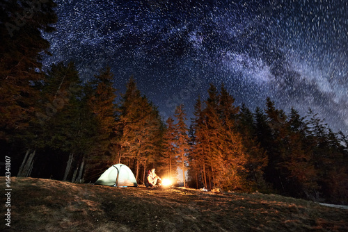 Male tourist enjoying in his camp near the forest at night. Man sitting near campfire and tent under beautiful night sky full of stars and milky way. Astrophotography © anatoliy_gleb