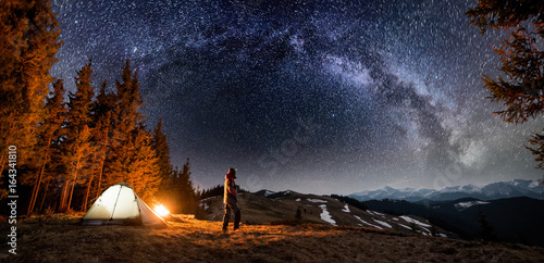 Male tourist have a rest in his camp near the forest at night. Man standing near campfire and tent under beautiful night sky full of stars and milky way, and enjoying night scene. Panoramic landscape