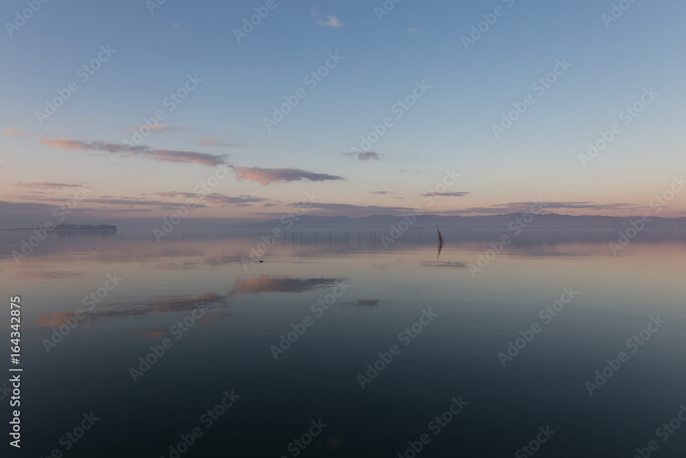 Fototapeta premium A symmetric photo of a lake, with a town and clouds reflections on water, soft and warm colors, and a water bird and wooden poles in the foreground