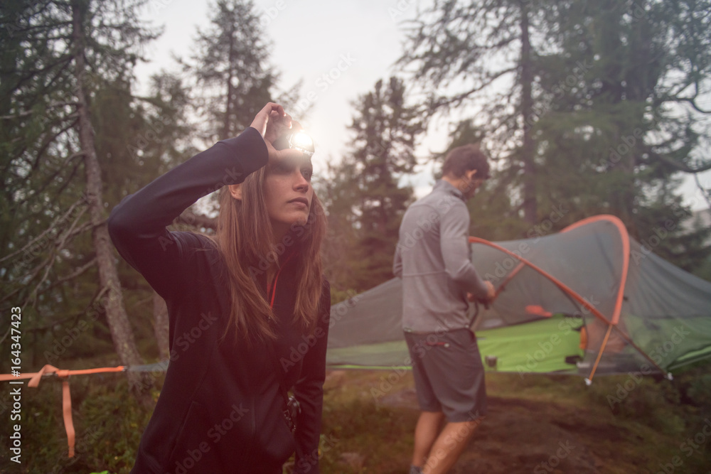 Woman turning on headlamp flashlight near hanging tent camping. Group of friends people summer adventure journey in mountain nature outdoors. Travel exploring Alps, Dolomites, Italy. - obrazy, fototapety, plakaty 