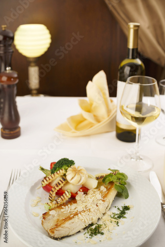 Hake fillet with grilled vegetables arranged on a plate, Wine bottle and wineglass in background, Traditional dish in elegant setting, Selective focus with soft light