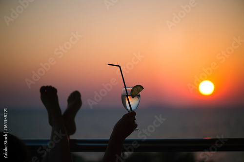 Female legs and glass with cocktail in the hand on sunset background.