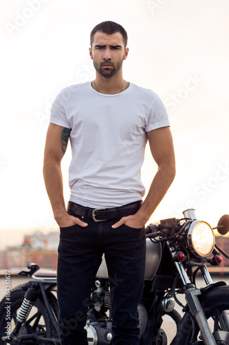 Close up of a handsome rider man in white blank t-shirt look to camera near classic style cafe racer motorcycle at sunset. Bike custom made in vintage garage. Brutal urban lifestyle. Outdoor portrait.