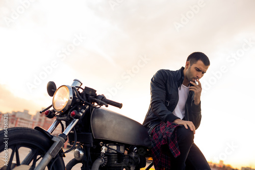 Handsome rider man with beard and mustache in biker jacket smoking, thinking and sit on classic style cafe racer motorbike at sunset. Bike custom made in vintage garage. Brutal fun urban lifestyle.