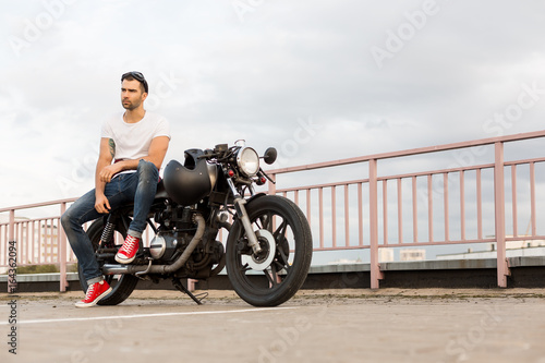 Rider guy with beard and mustache in red sneakers sit on classic style biker cafe racer motorcycle with black glasses and look camera. Bike custom made in vintage garage. Brutal fun urban lifestyle.