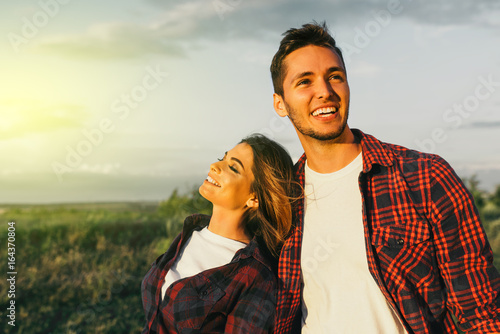 Carefree stylish young Caucasian male and female enjoying sunset in meadow while spending summer vacations talking, laughing and having fun. Happy couple enjoy honeymoon in mountain.