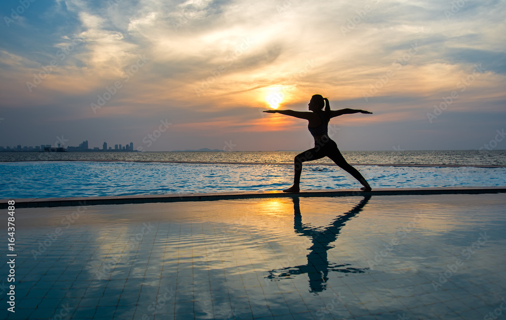 Silhouette young woman practicing yoga on swimming pool and the beach at sunset.  Healthy Concept..