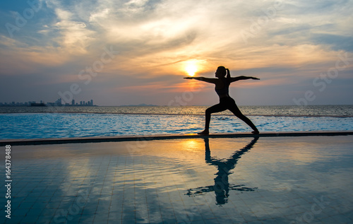 Silhouette young woman practicing yoga on swimming pool and the beach at sunset. Healthy Concept..