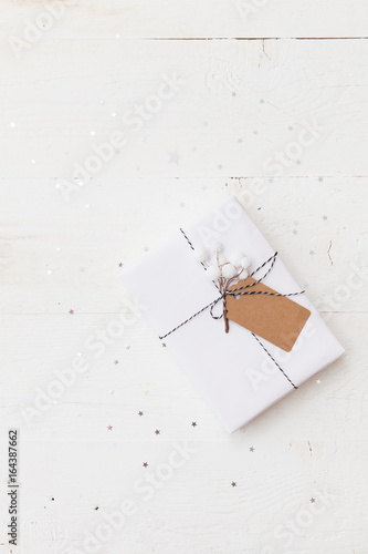 Top view on nice Christmas gift wrapped in white gift paper, Christmas tree decorations on white wooden background with sparkling stars. New Year, holidays and celebration concept