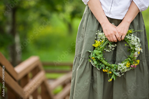 Woman with wild flowers wreath in her hands walking in the park. Girl standing at the wooden stairway on a sunny summer day. Outdoors. Summer and lifestyle concept