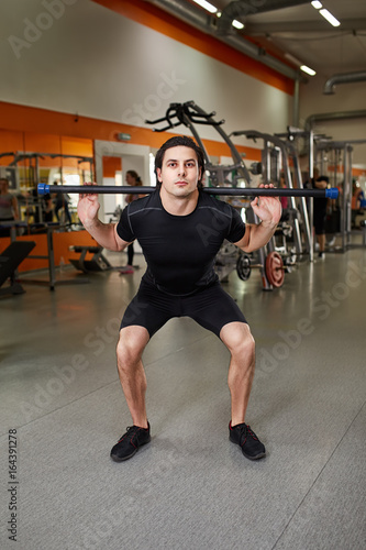 Young sporty man in the black sportwear with barbell doing squats in gym.