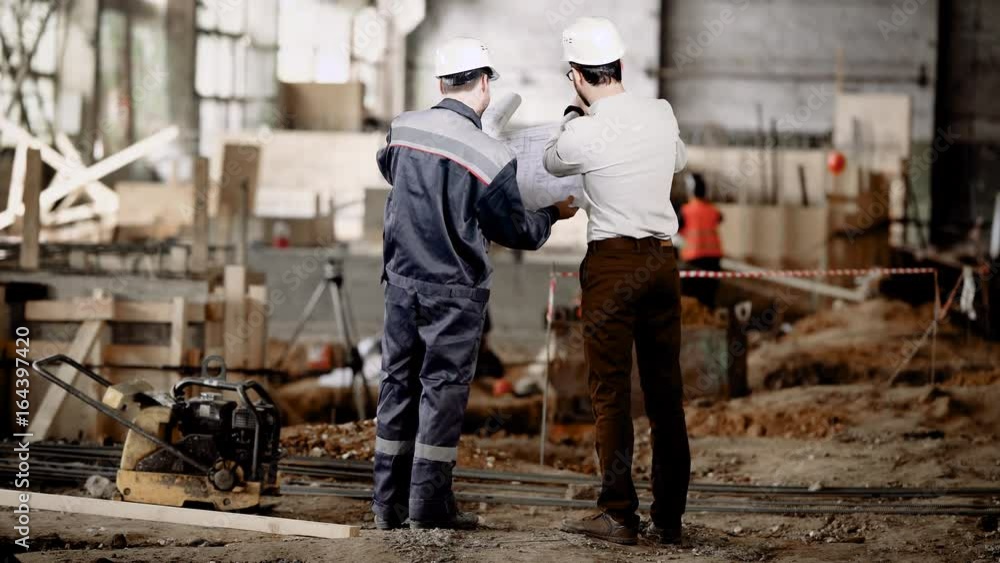 Back view of two men standing together in construction area and looking at shop drawings of future structure. Foreman in shirt and trousers is discussing with worker about details of building-up.