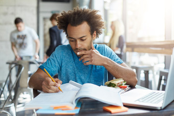 Indoor shot of dark-skinned male student with African hairstyle wearing blue casual T-shirt sitting at university canteen eatting hamburger reading attentively book underline necessary information