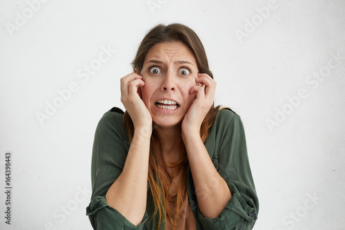 Concerned woman with bugged dark eyes keeping hands on cheeks looking desperately being scared of something horrorful. Shocked female looking terrified. Facial expression and emotions concept photo