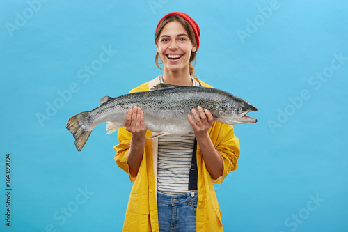 Glad fisher woman in yellow anorak and jeand overalls holding huge fish rejoicing to catch it demonstrating her work while standing over blue background. People, hobby, recreation and fishing