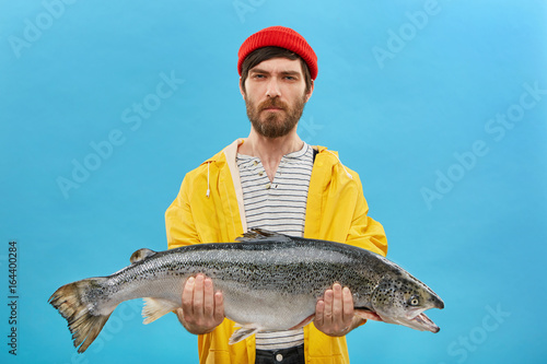 Young unshaven fisherman spending many days on lake cathcing fish holding his big catch in hands looking tired and serious. Attractive young male wearing yellow raincoat holding big fish in hands