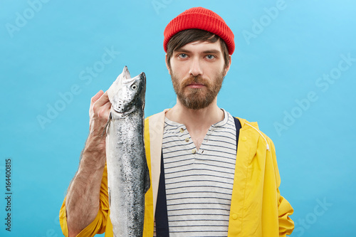 Indoor shot of handsome young European fisherman with beard showing his catch after fishing excursion. Confident male wearing sailor shirt, raincoat and hat posing in studio with big sea fish