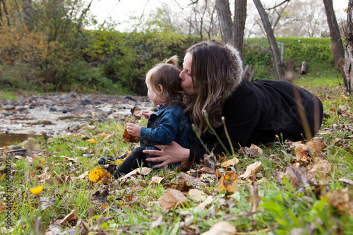 mother sits behind her small daughter at the park by a creek, kissing back of her head 