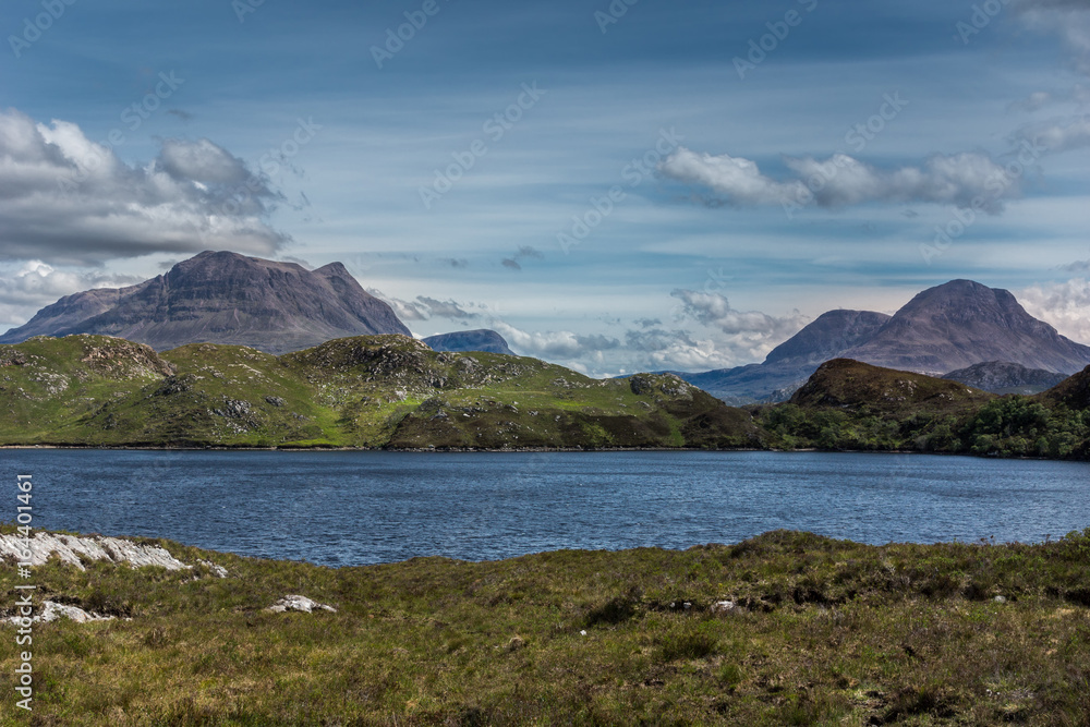 Assynt Peninsula, Scotland - June 7, 2012: multiple brown mountain peaks under blue skies with gray clouds fronted by green wild rough slopes east of Loch Buine Moire. Lake waters in front.