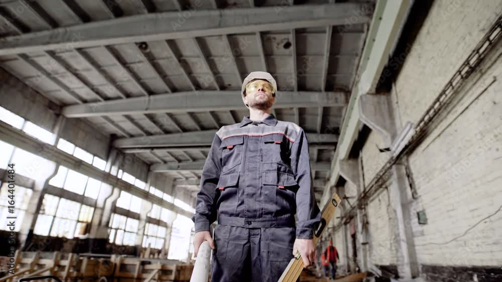 Young foreman standing in the building plant with welding in the background. Worker is holding construction plan and spirit level, checking process of construction and looking over working area. Young foreman standing in the building plant with welding in the background. Worker is holding construction plan and spirit level, checking process of construction and looking over working area.