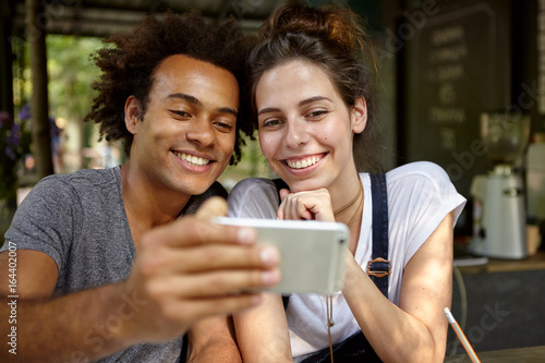 Portrait of black Afro American male and his European girlfriend sitting close to each other at cozy restaurant looking with smiles in camera of their smart phone making selfie. People, lifestyle