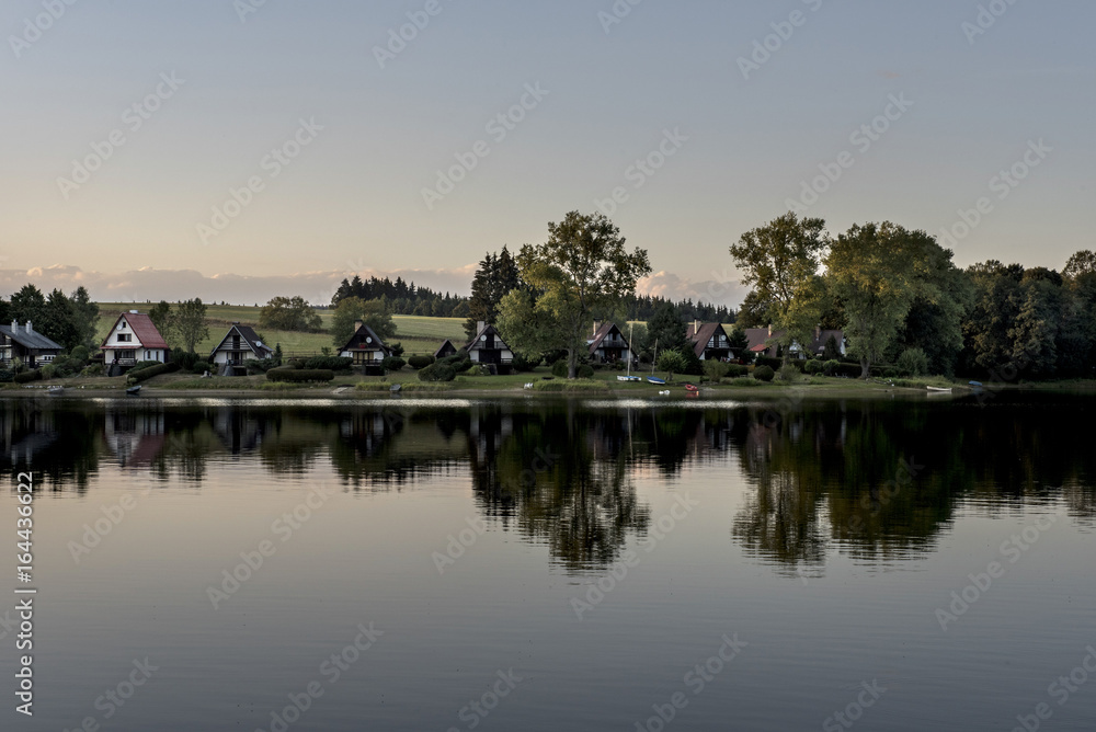 Small houses on the shore of the Lipno dam reflecting in the water at sunset around the woods