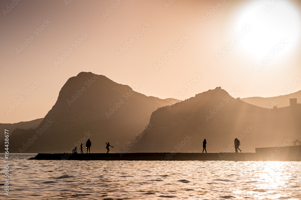 Silhouettes of people and rocks against the setting golden sun on the black sea. Shaft of lights make Silhouette. Shadow at sunset. Black sea beach in Sudak, Crimea.