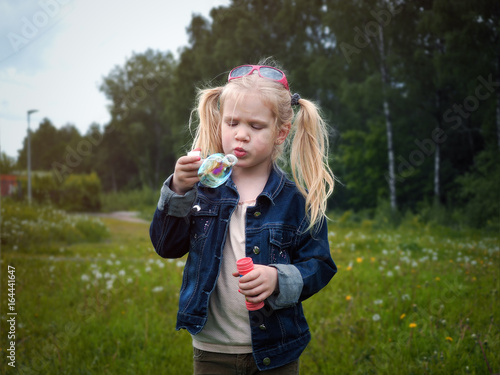Girl inflates soap bubble. Portrait of a child. Long blonde hair, beautiful face. Nature, field, green, grass, forest