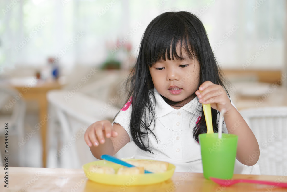 Asian children cute or kid girl enjoy eating dessert and bread delicious with saliva on lips and colorful container or utensil plastic for baby on wood table in cafe restaurant for breakfast and lunch Asian children cute or kid girl enjoy eating dessert and bread delicious with saliva on lips and colorful container or utensil plastic for baby on wood table in cafe restaurant for breakfast and lunch