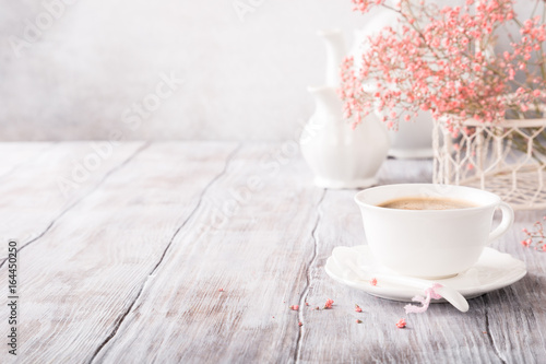 White cup of coffee with pink flowers on old white wooden background with copy space.
