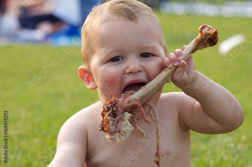 A baby boy is eating a large turkey leg. He is really enjoying his picnic 