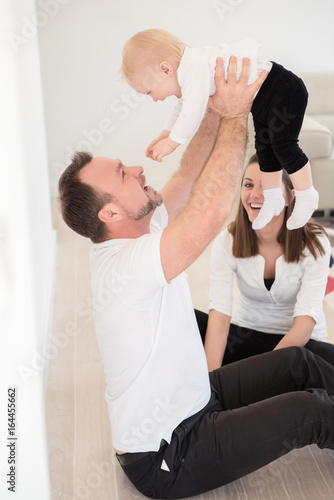 Happy family of three is enjoying at home. Parents and their beautiful baby girl sitting on the ground and playing. Father raise up his daughter. Family values. Leisure together.