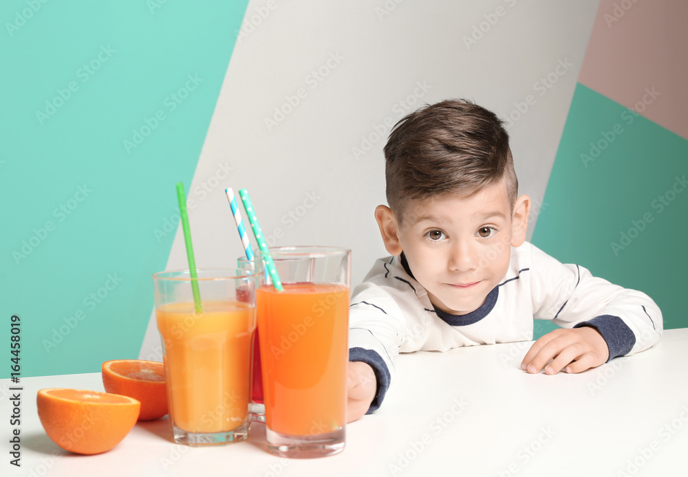 Cute little boy making long arm for glass of juice while sitting at table, on color background Cute little boy making long arm for glass of juice while sitting at table, on color background