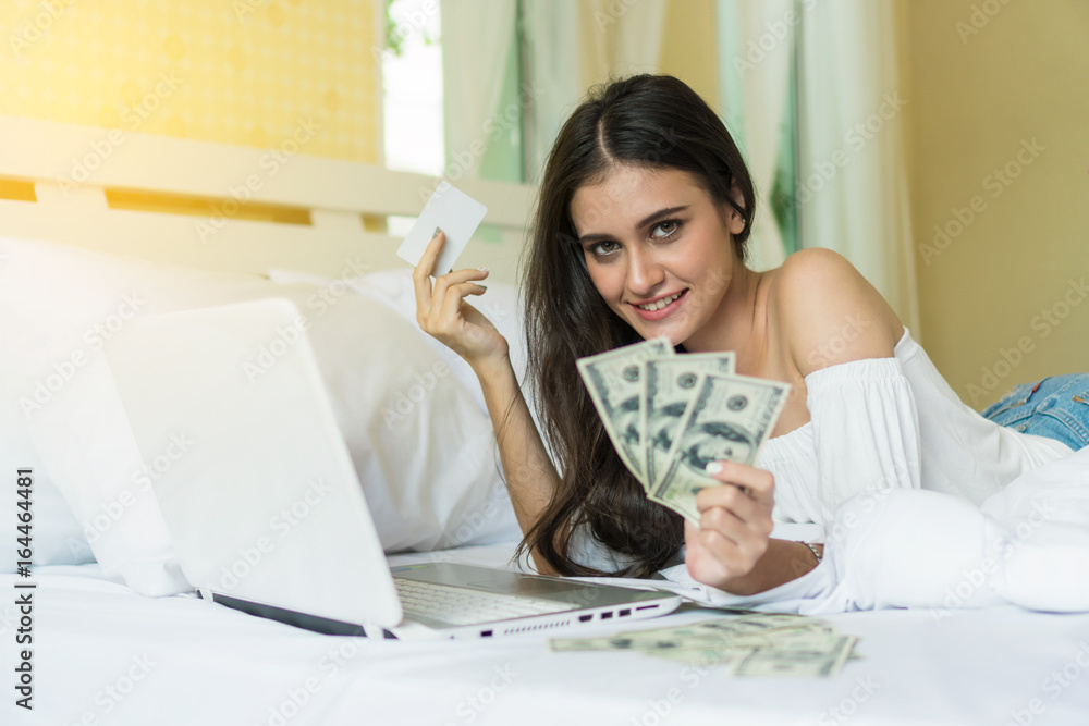 Beautiful smiling young woman lying in bed holding dollar and credit card with a laptop in front to shop online. Beautiful smiling young woman lying in bed holding dollar and credit card with a laptop in front to shop online.
