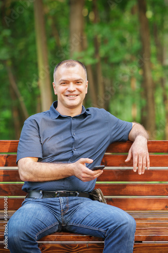 adult man around 35-40 is sitting with smile and phone on the bench atsummer park