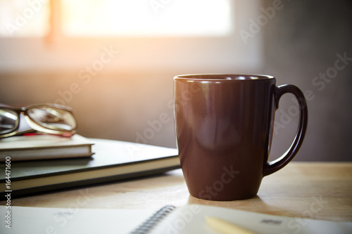 Morning coffee on the desk with sunlight from window, 
Coffee mug on the table with the blurred of notebook and glasses