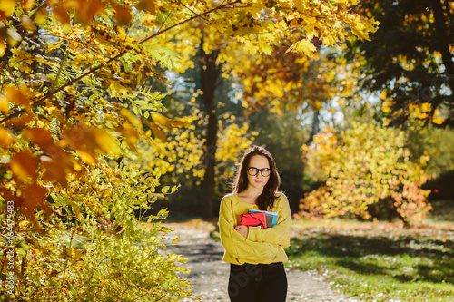 Portrait of pretty student girl with books in a autumn park, adult schoolgirl enjoying education, beautiful female wearing with yellow sweater , scarf and glasses, back to school