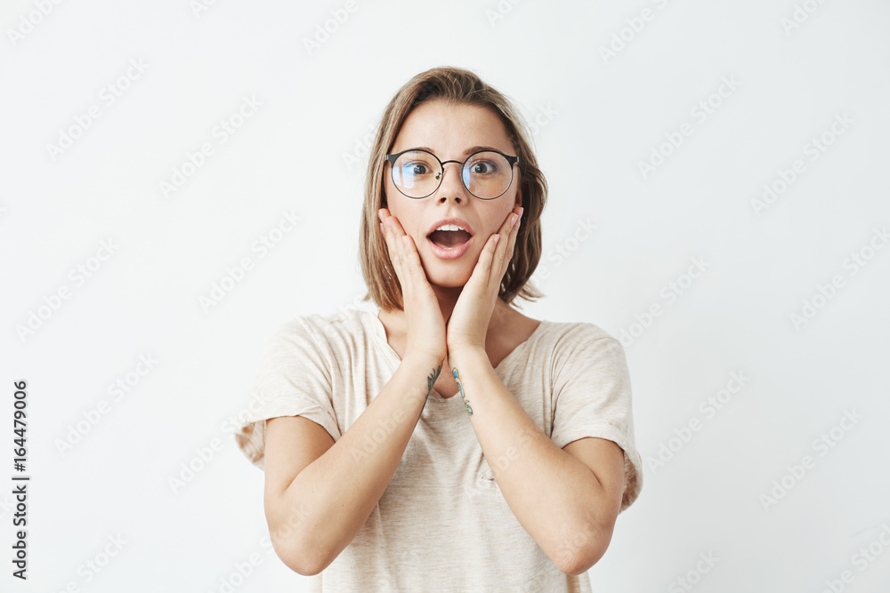 Surprised young beautiful girl in glasses looking at camera with opened mouth and hands on cheeks over white background Surprised young beautiful girl in glasses looking at camera with opened mouth and hands on cheeks over white background